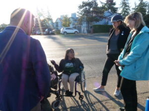 A group of people on the sidewalk talking to someone in a wheelchair who is pointing towards something on the street.